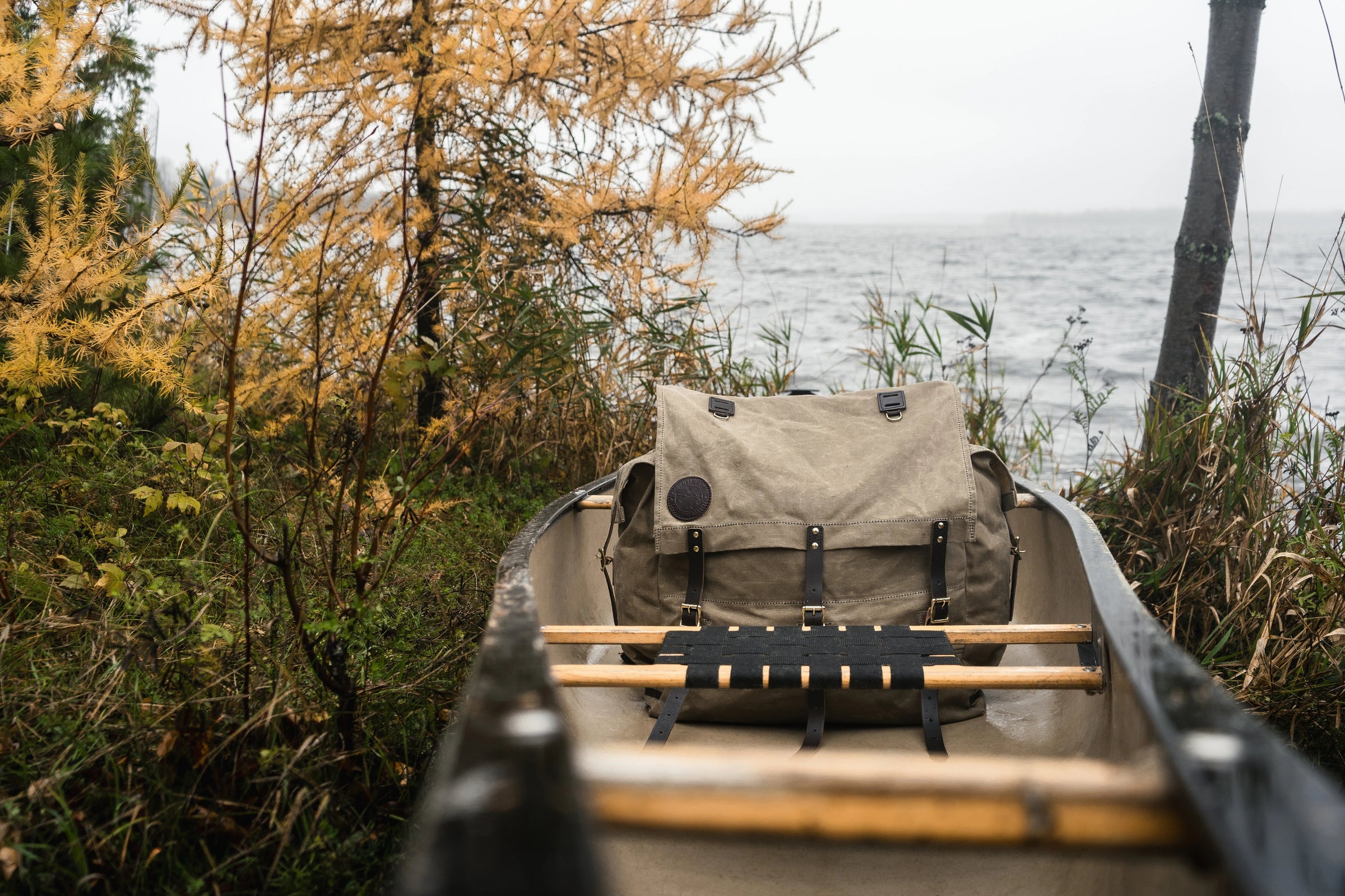 A Duluth Pack sitting in a canoe on the edge of a windy lake. The canoe is surrounded by trees.