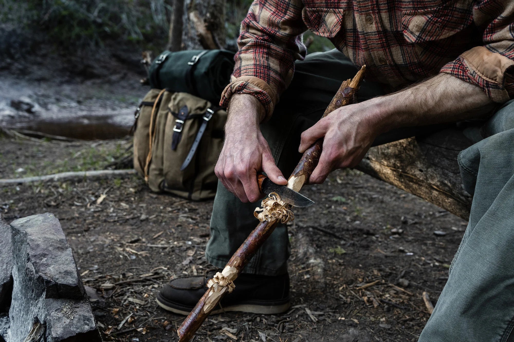 A man wittling a piece of wood with a Helle Knive. There is a Duluth Pack Bushcrafter Pack in the background. 