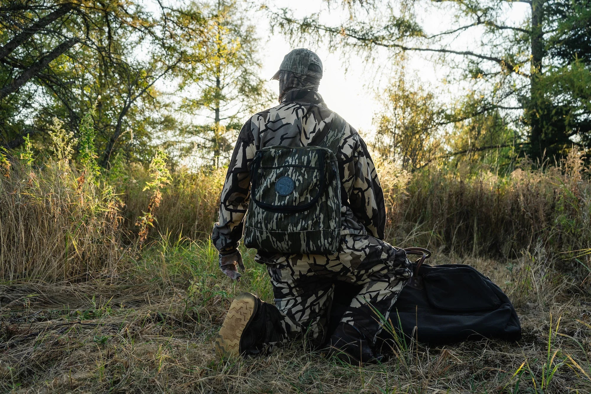 A man wearing hunting mossy oak camo with a Duluth Pack Blind Bag with a forest in the background. 