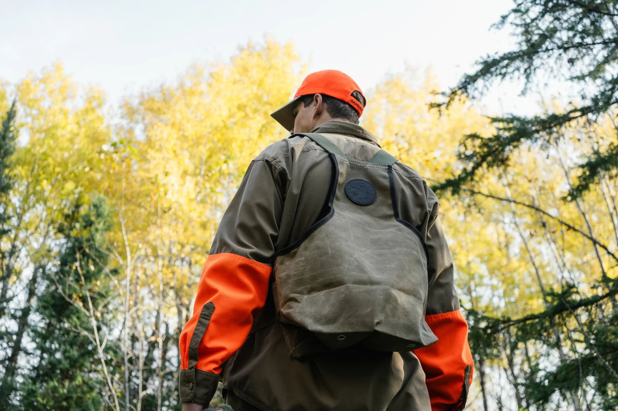 A man wearing pheasant hunting clothes and wearing a Waxed Khaki Duluth Pack Upland Game Vest. There are fall color trees in the background.