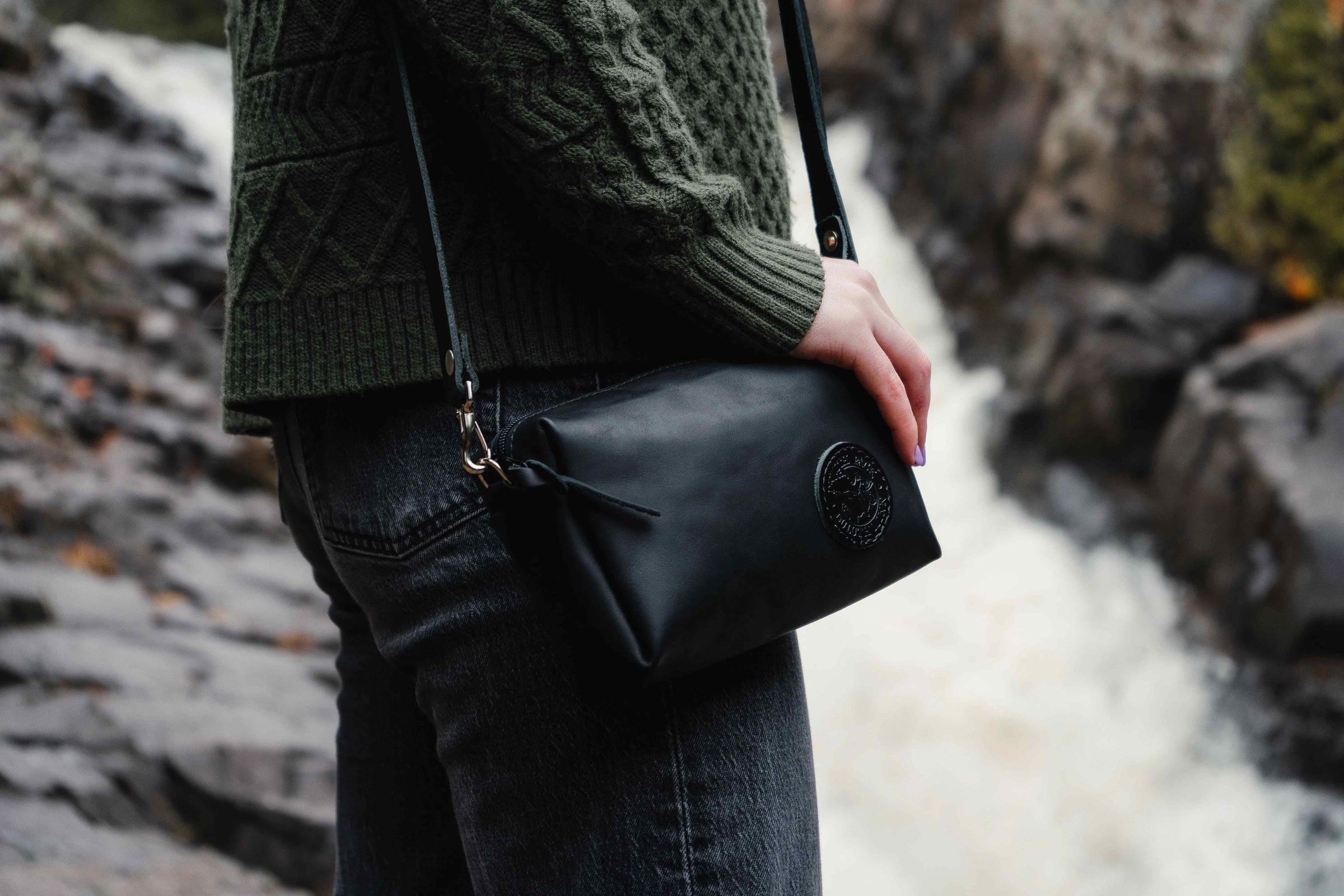 A girl wearing a black Duluth Pack black leather Riverwalk purse with a waterfall in the background. 