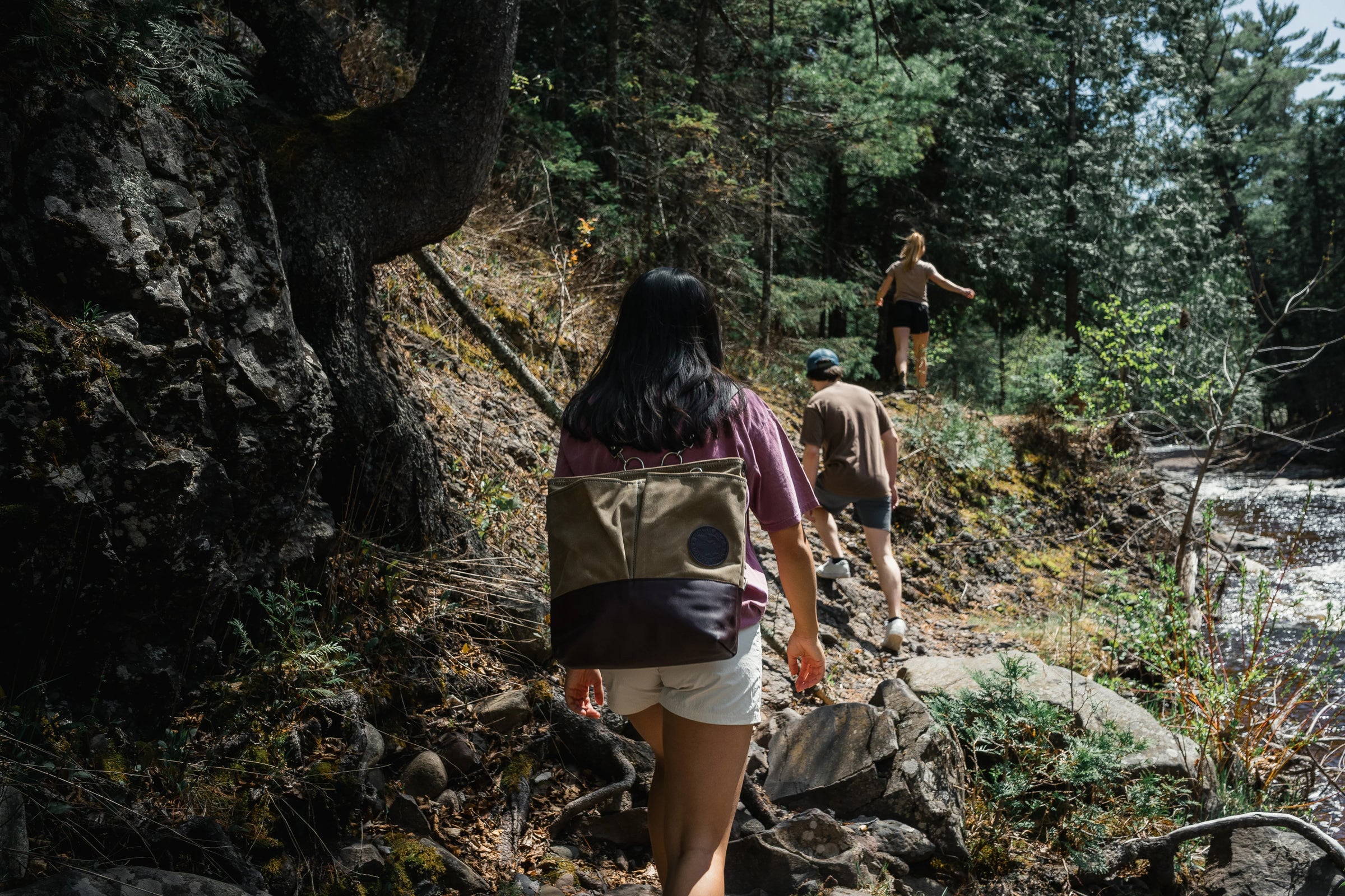 Woman wearing a convertible jet setter tote walking through the woods with another guy in the background.
