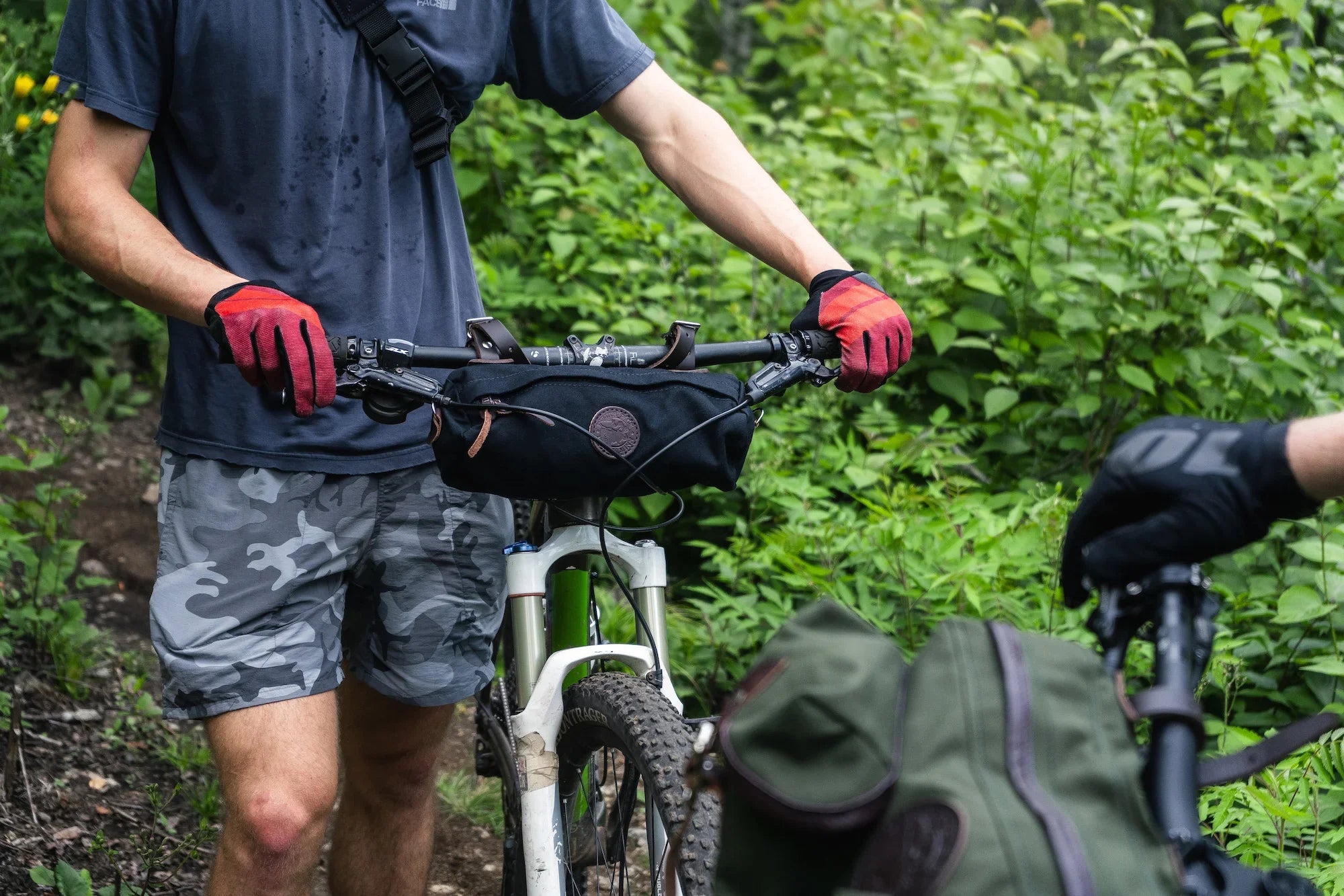 A man standing with a mountain bike. On the mountain bike is a Duluth Pack Candy Bar Bike Bag with a green forest in the background. 
