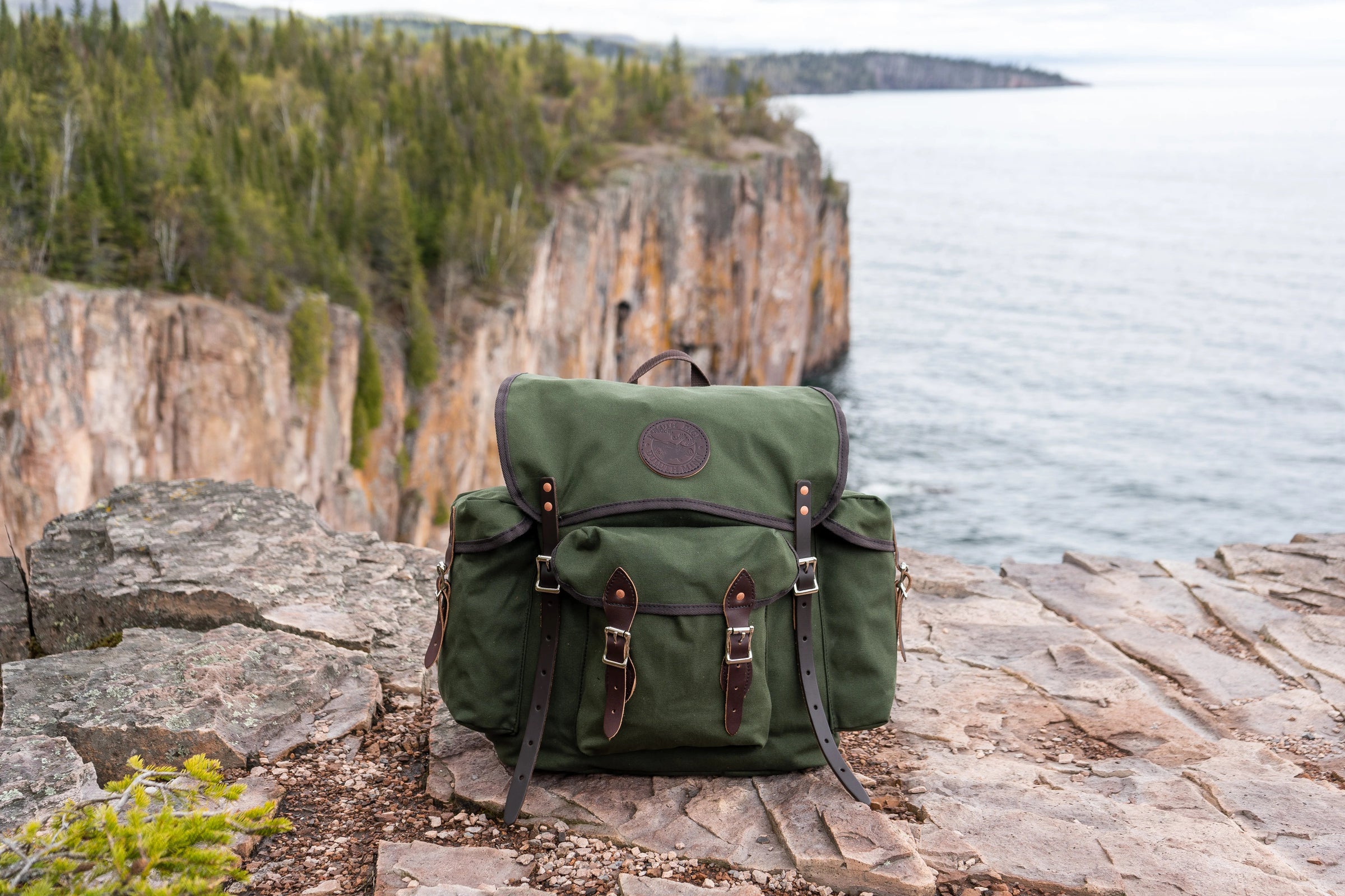 A Duluth Pack Wanderer pack in Olive Drab sitting on a cliff with another large cliff in the background. 