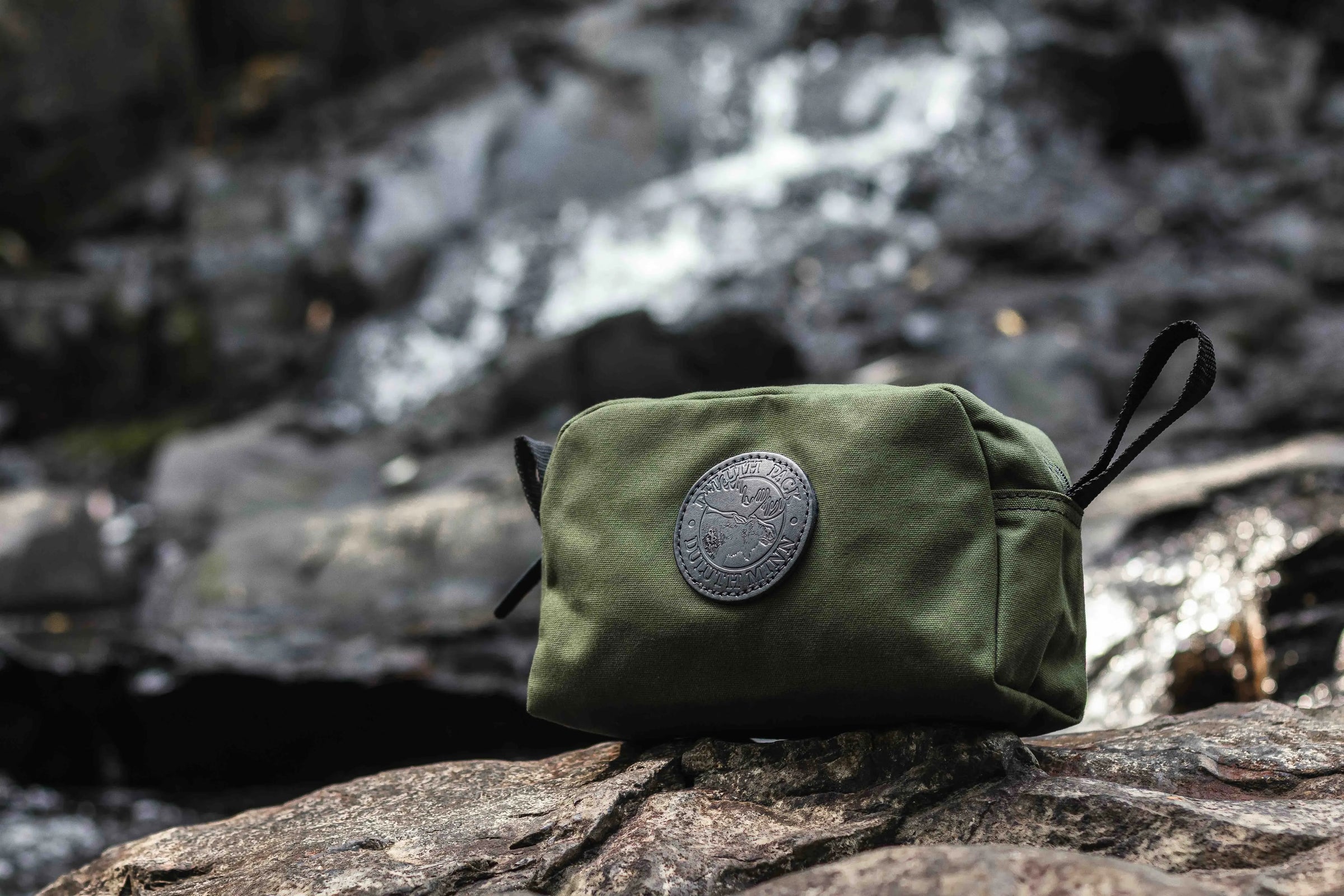 A olive drab/green Duluth Pack Grab-N-Go sitting on a rock with a waterfall in the background. 