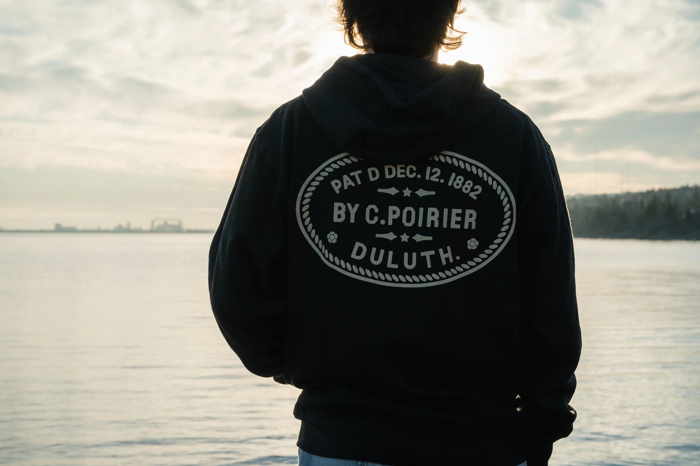 Man with his back to the camera facing a still lake. The man is wearing a Duluth Pack sweatshirt. 