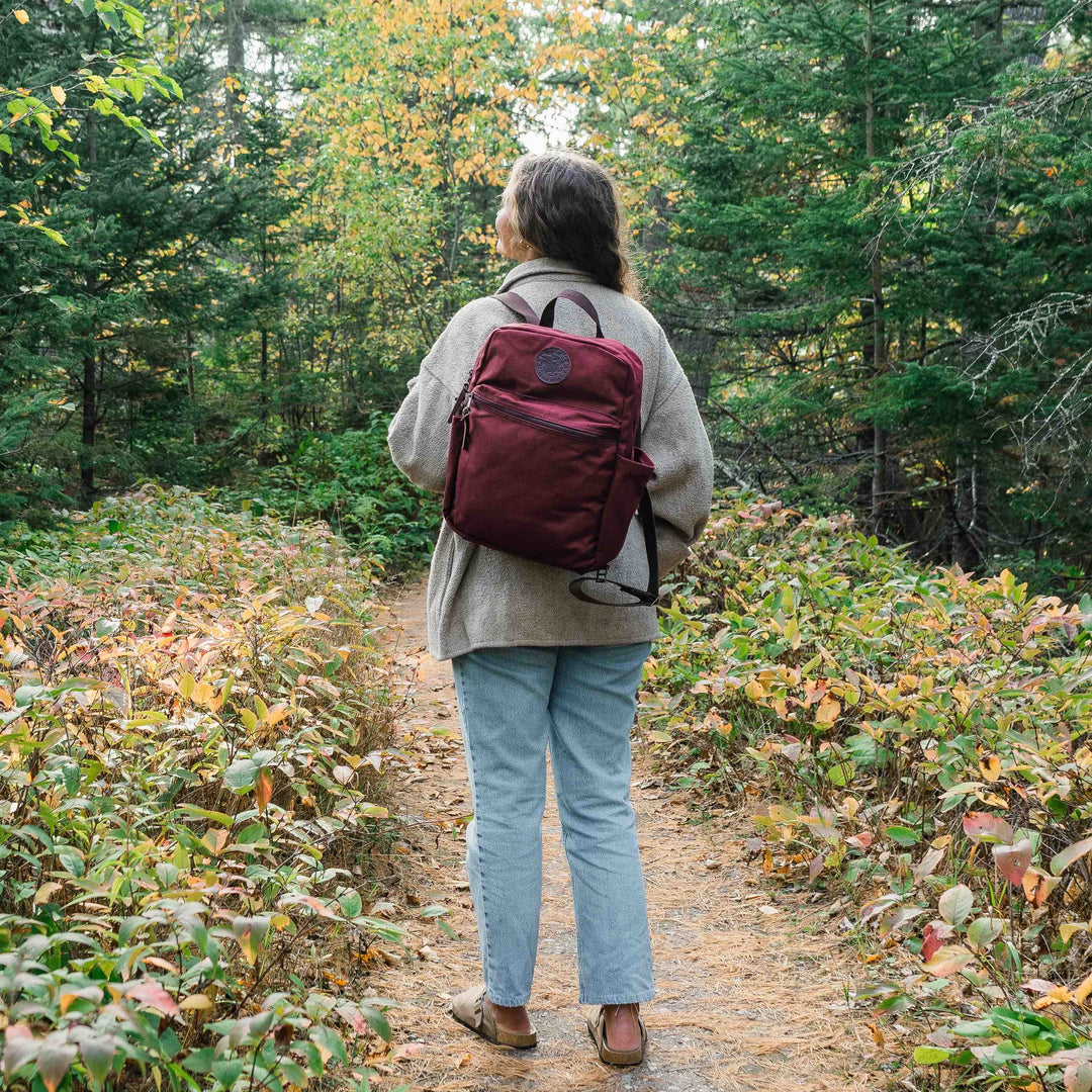 A woman wearing a burgundy Standard Backpack by Duluth Pack with a forest in the background