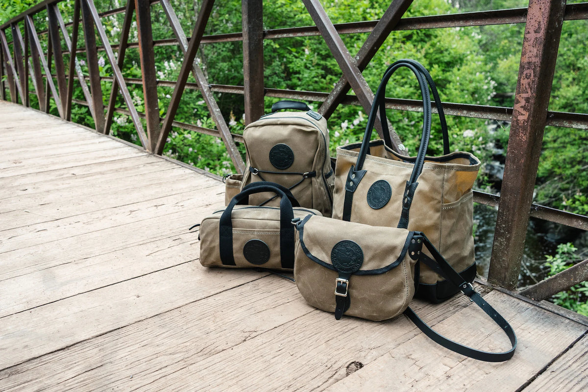 Duluth Pack Lake Ontario collection featuring a group of waxed khaki and black bags sitting on a bridge with a forest in the background. 